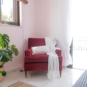 A red chair with white cushions and a white blanket, a potted plant, and a view of a balcony with a railing. - Pefkides Aegina Apartments (Aegina)