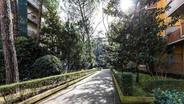 A pathway lined with trees and bushes, leading towards a building with balconies.
