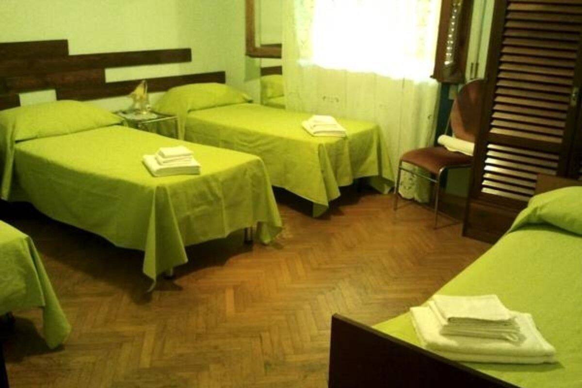 Une chambre avec trois lits simples, chacun recouvert d’une couverture verte et agrémenté d’une petite table sur laquelle se trouvent un livre et une lampe. La chambre est dotée d’un plancher en bois et d’une fenêtre avec des rideaux.
