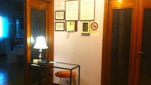A hotel room with a wooden door, a glass table with a lamp, a chair, and several framed documents on the wall.