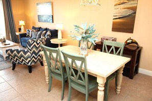 Un comedor con una mesa de madera y sillas verdes, una mesita auxiliar con una lámpara, y un jarrón decorativo con una planta.