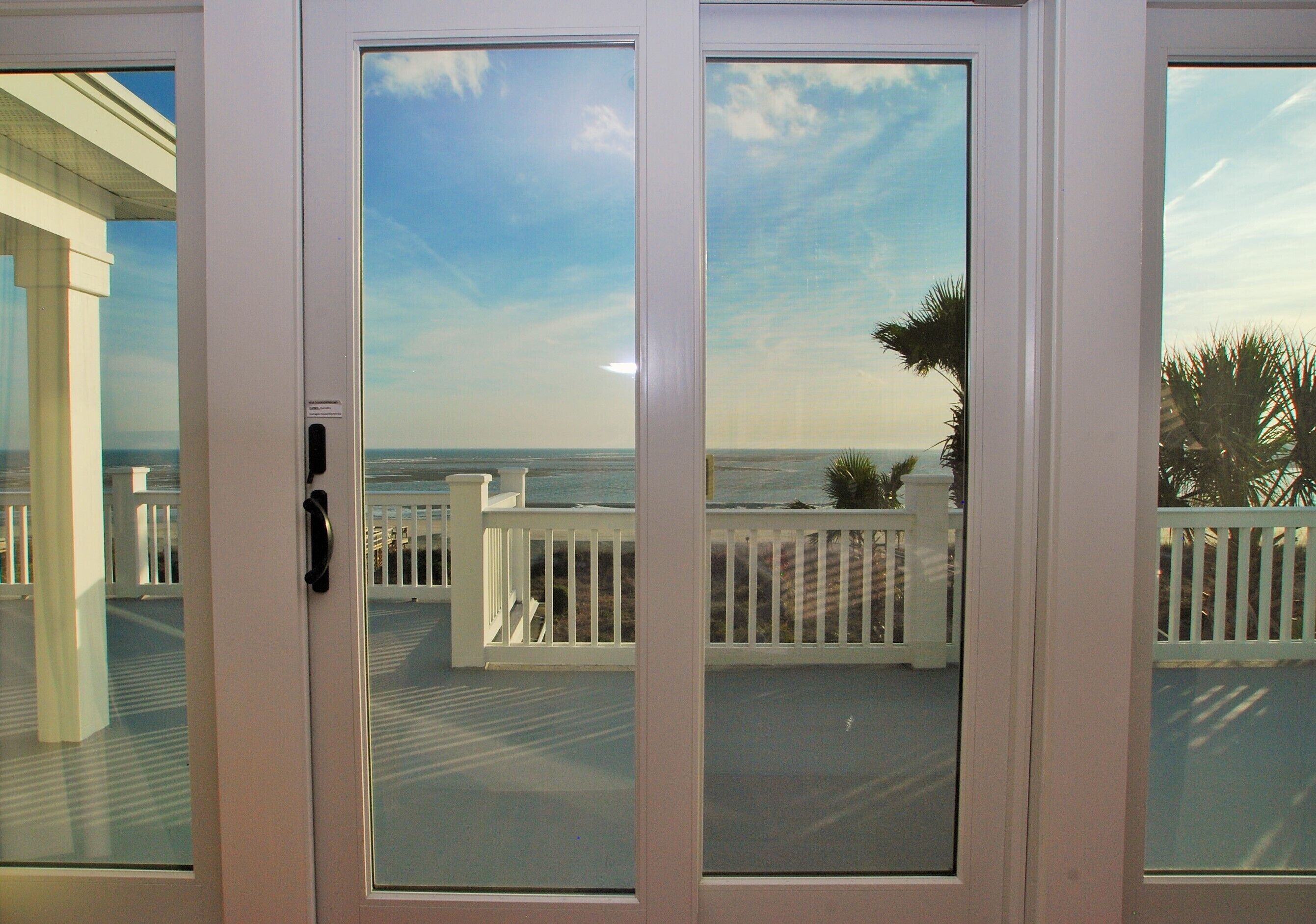 A view through a glass door to a balcony with a white railing, overlooking a beach and ocean.