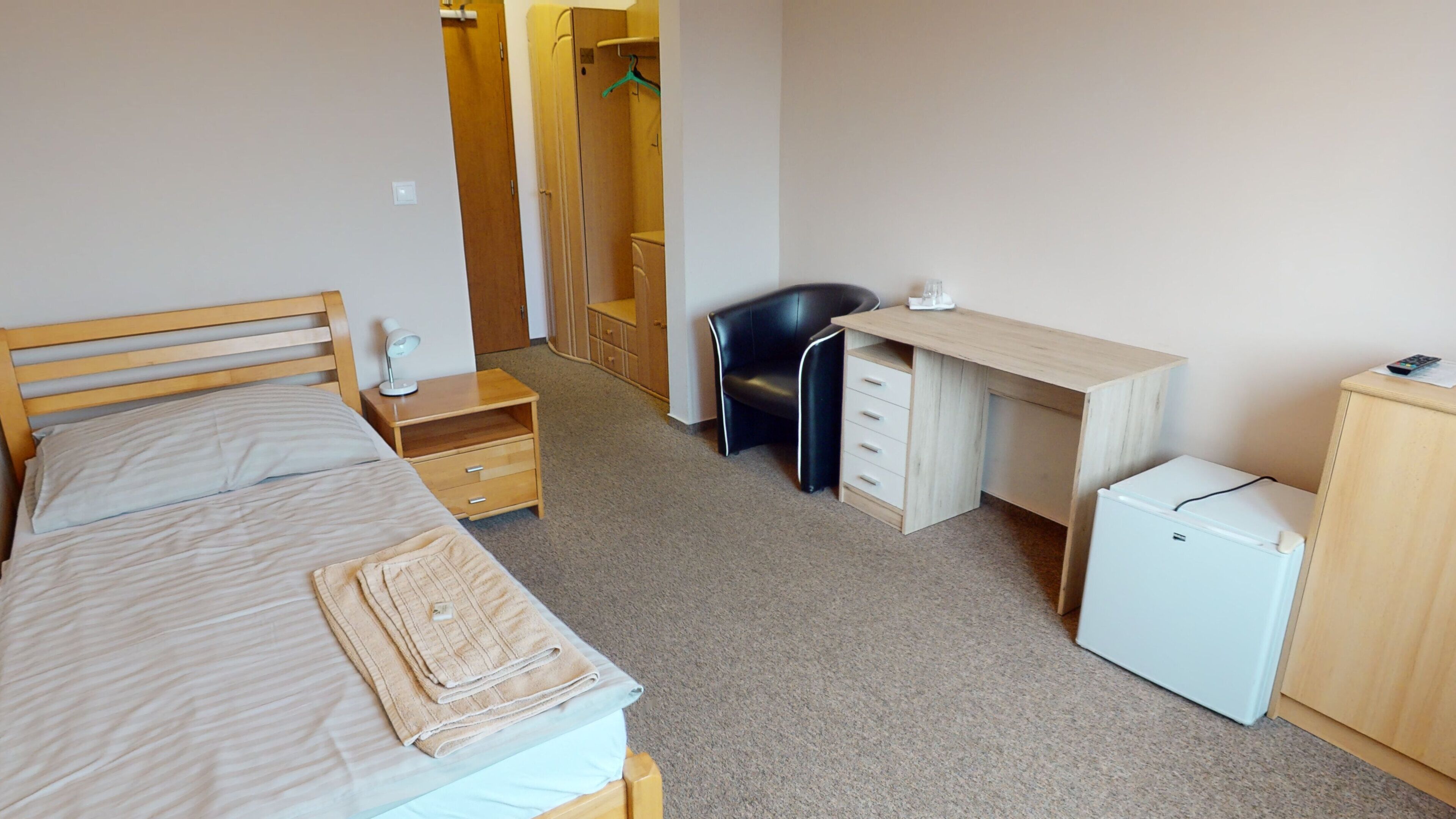 A bedroom with a bed, bedside table, lamp, desk, chair, and a small refrigerator.