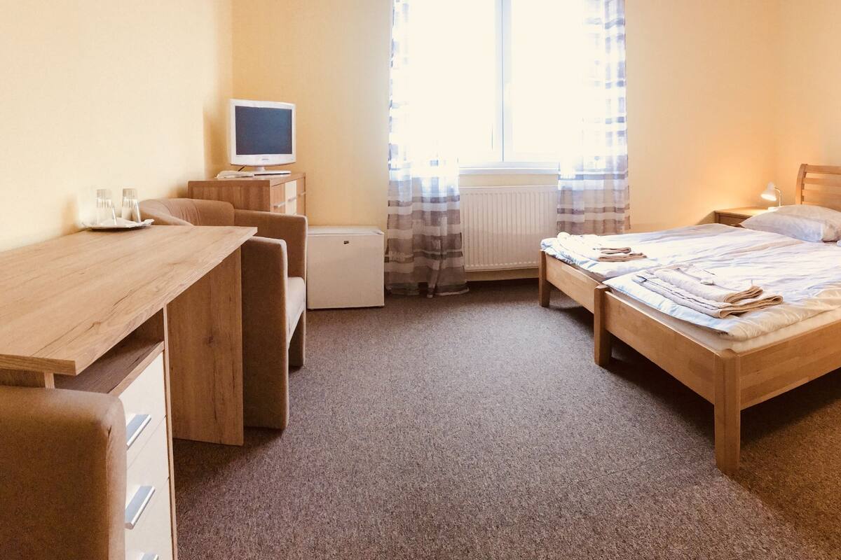 A bedroom with a bed, bedside tables, a desk, a chair, a radiator, and a window with curtains.