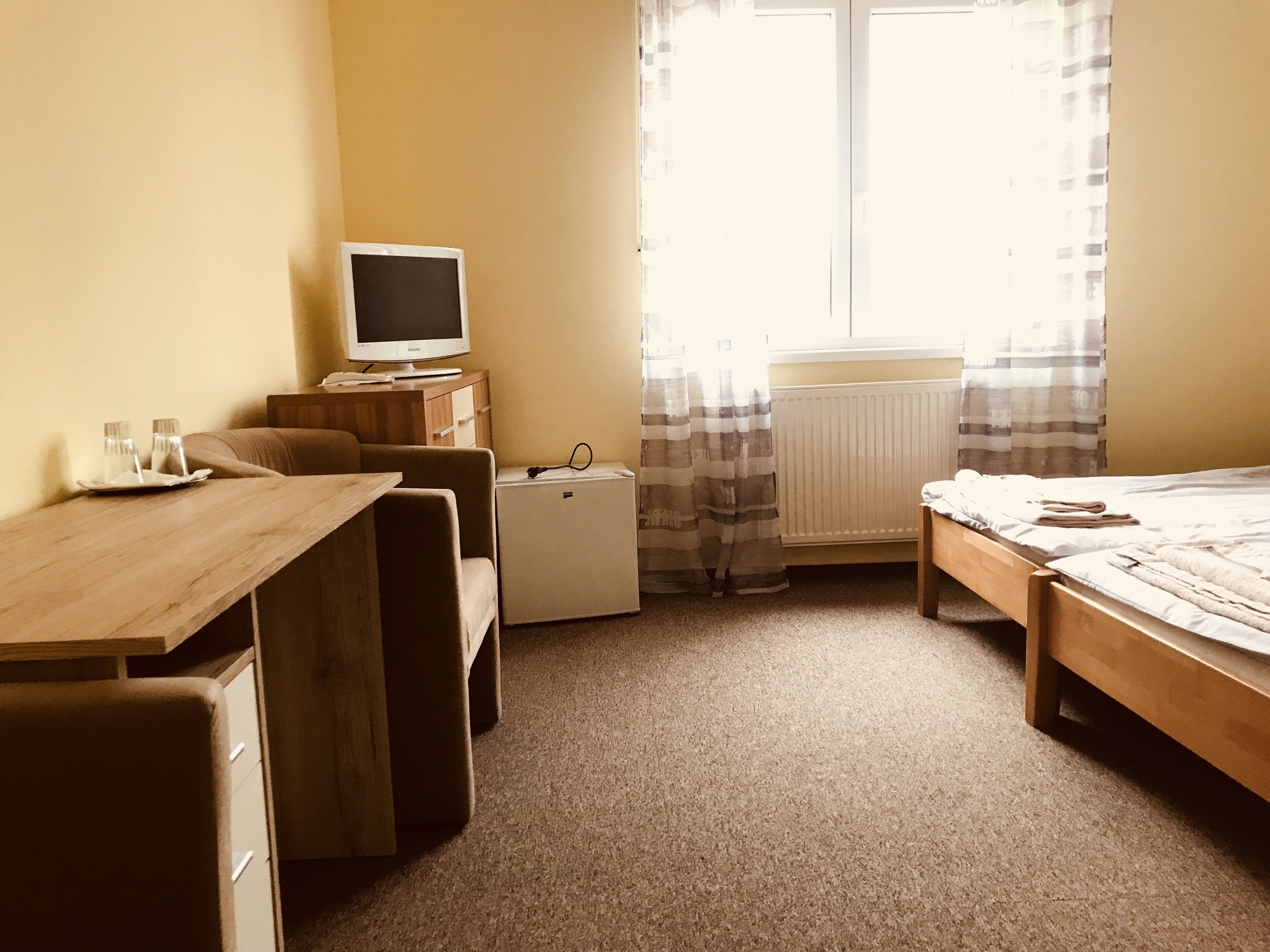 Een compacte hotelkamer met een houten bureau, een kleine koelkast, een bed met een witte dekbedovertrek en een raam met gordijnen.