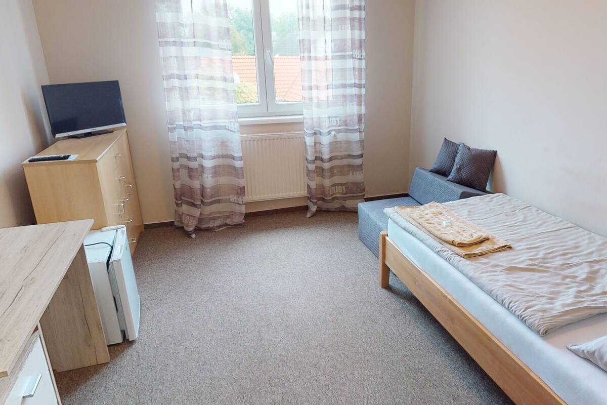 A small, tidy room with a bed, a desk, a television, and a window with curtains.