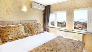 Una habitación de hotel con una cama, ropa de cama estampada con motivos florales, vistas de la ciudad a través de amplias ventanas y un equipo de aire acondicionado.