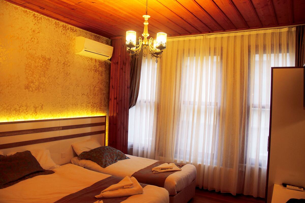 A hotel room with two beds, a chandelier, an air conditioning unit, and a large window with sheer curtains.
