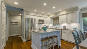 A modern kitchen with white cabinets, stainless steel appliances, and a central island with stools. - Coastal Calm- Luxury in the Lowcountry (Kiawah Island)