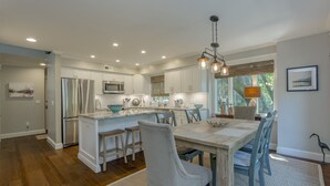 A modern kitchen with white cabinets, stainless steel appliances, and a wooden dining table with chairs. - Coastal Calm- Luxury in the Lowcountry (Kiawah Island)