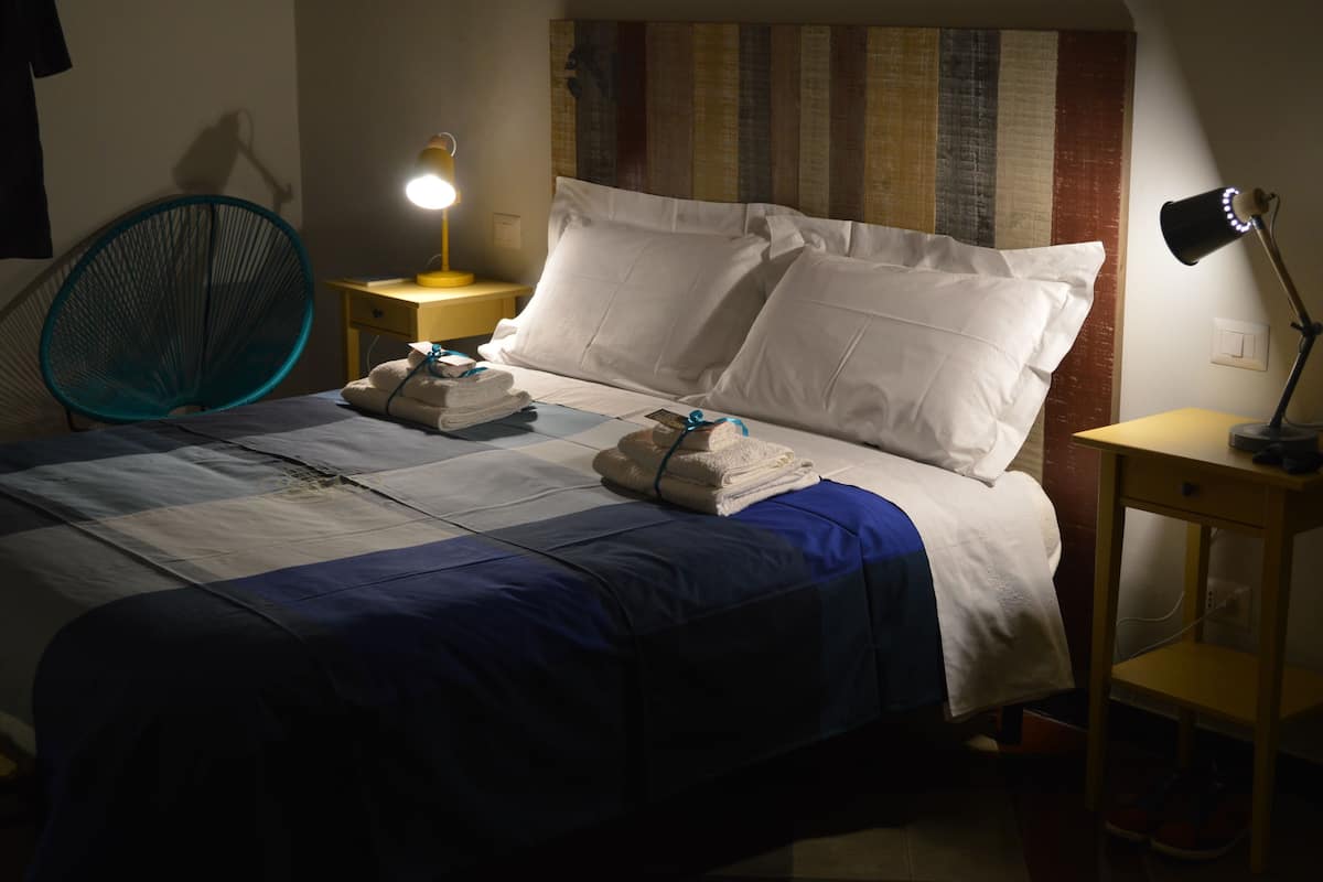A neatly made bed with white and blue bedding, a folded towel on the bed, a standing lamp, and a chair.