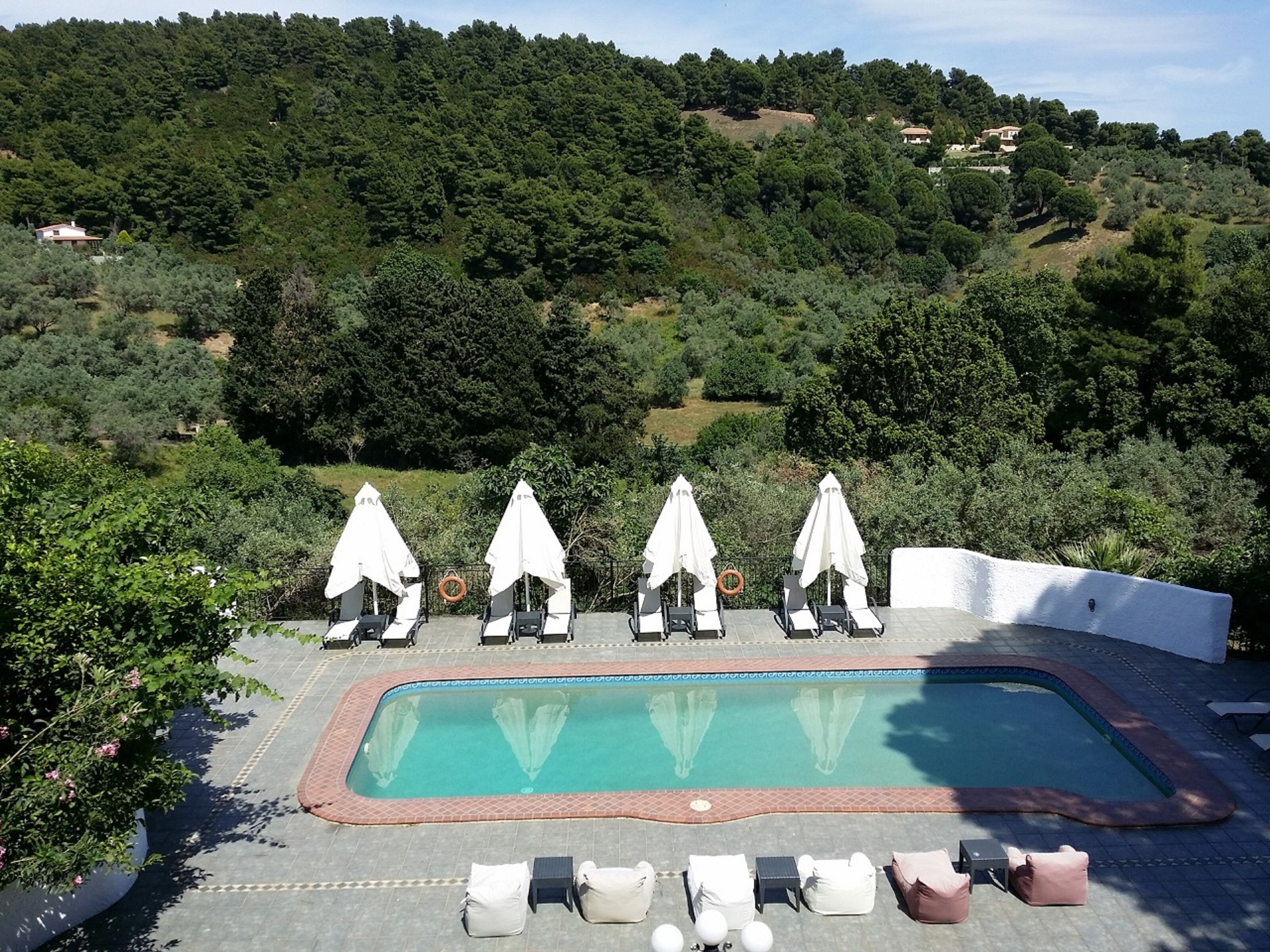A pool area with lounge chairs and umbrellas, surrounded by lush greenery and hills in the background.