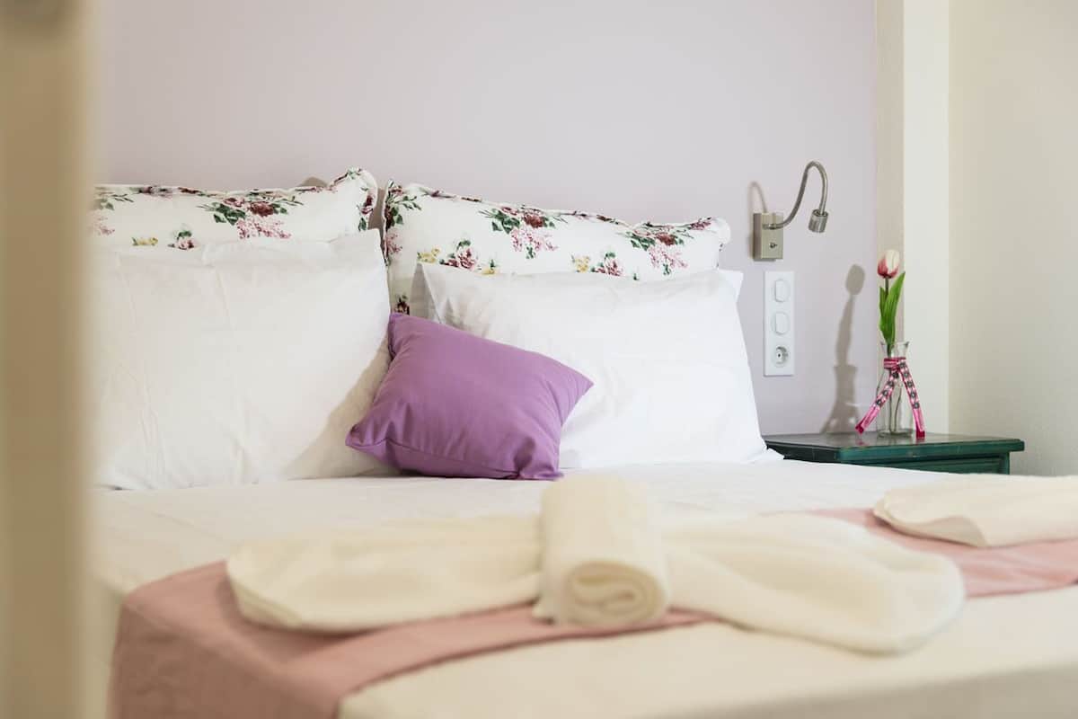 A neatly made bed with white and purple pillows, a green nightstand with a lamp, and a vase with flowers.
