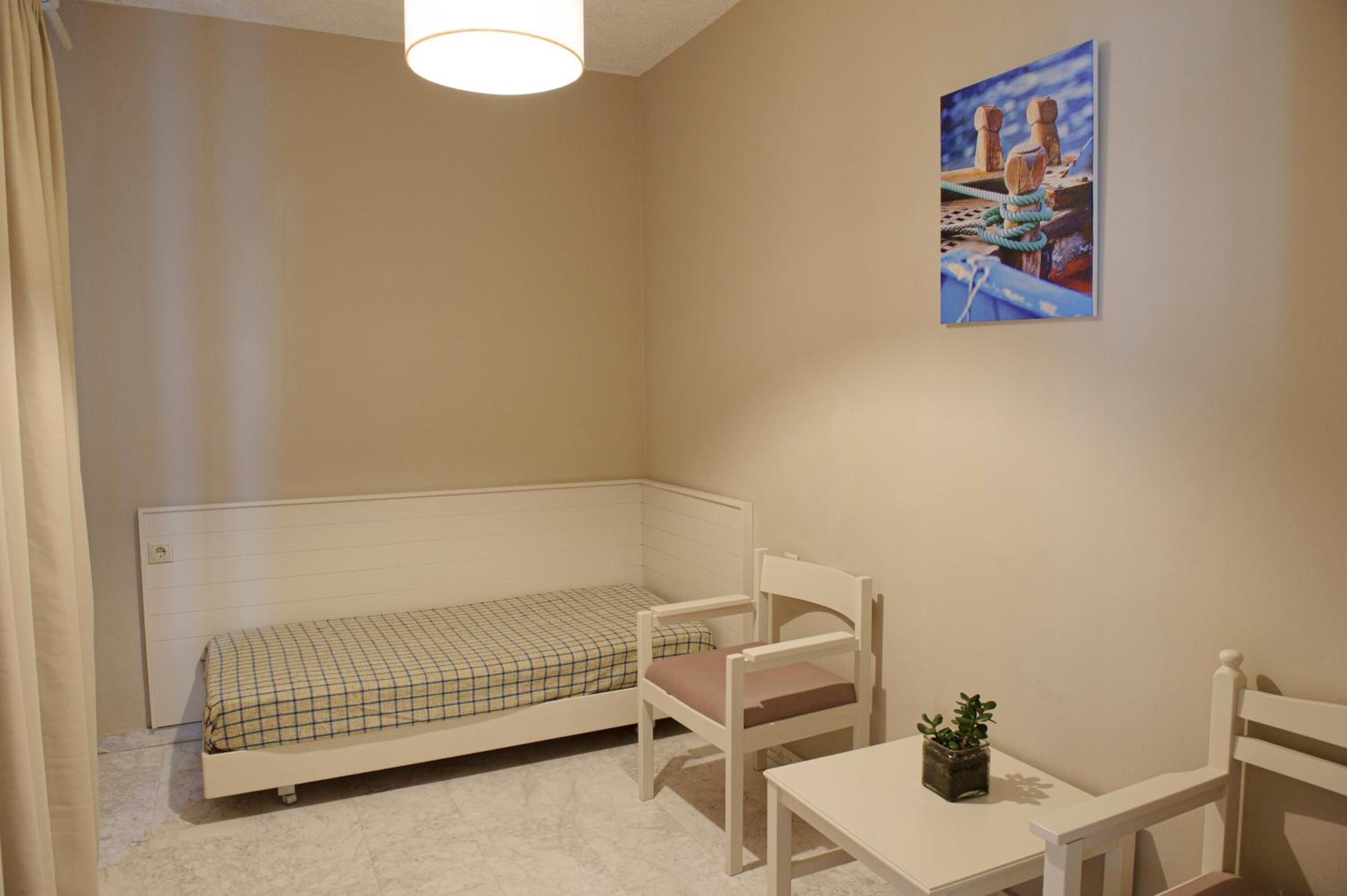 Ein kleines, ordentlich eingerichtetes Schlafzimmer mit einem Einzelbett, einem Sessel, einem Tisch und einem Wandbild.