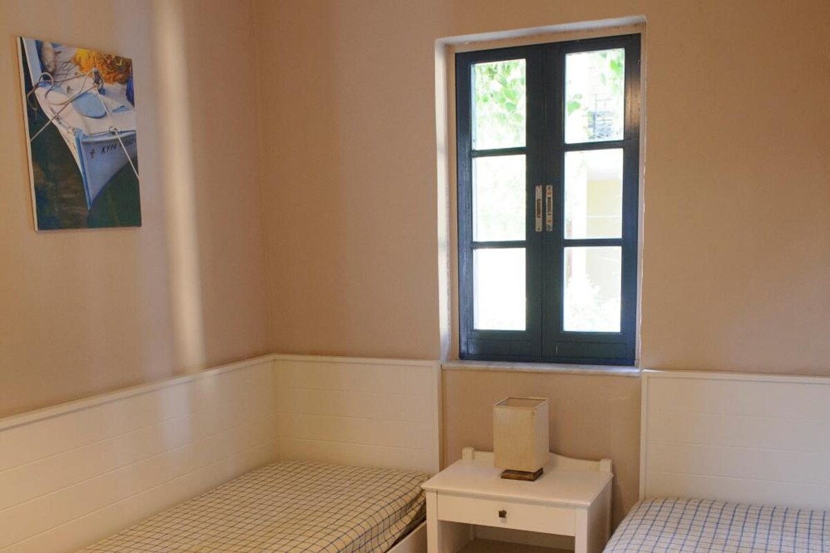 A small, clean room with two single beds, a window with a view, and a bedside table with a lamp.
