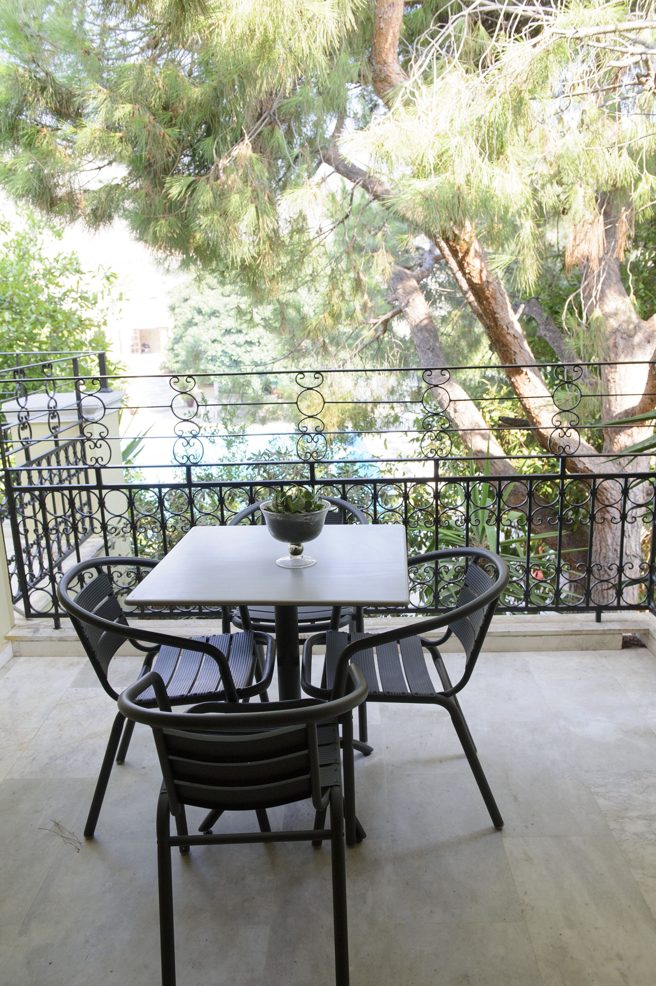 A balcony with a table and chairs, a potted plant, and a view of trees and a building.