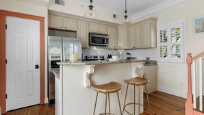 A kitchen with wooden cabinets, a stainless steel refrigerator, a microwave, and a bar area with stools. - Cozy Charleston Cottage | Brandy Crusta (Charleston)