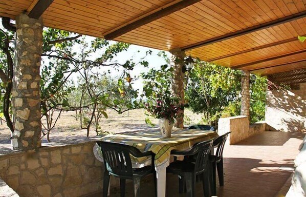 Un patio techado al aire libre con una mesa y sillas, rodeado de muros de piedra y vegetación.
