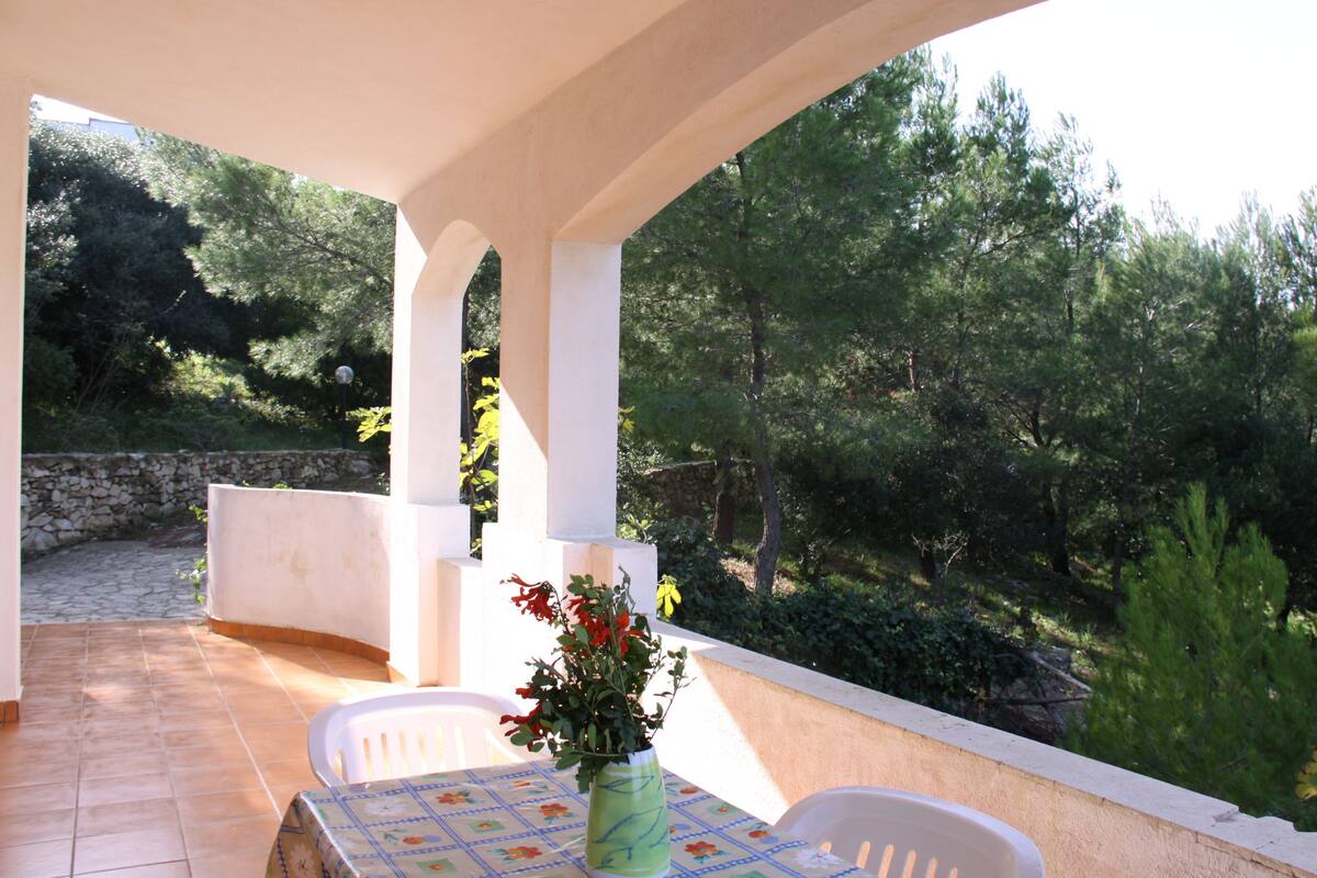 A covered patio with a table and chairs, a vase with flowers, and a view of a forested area.