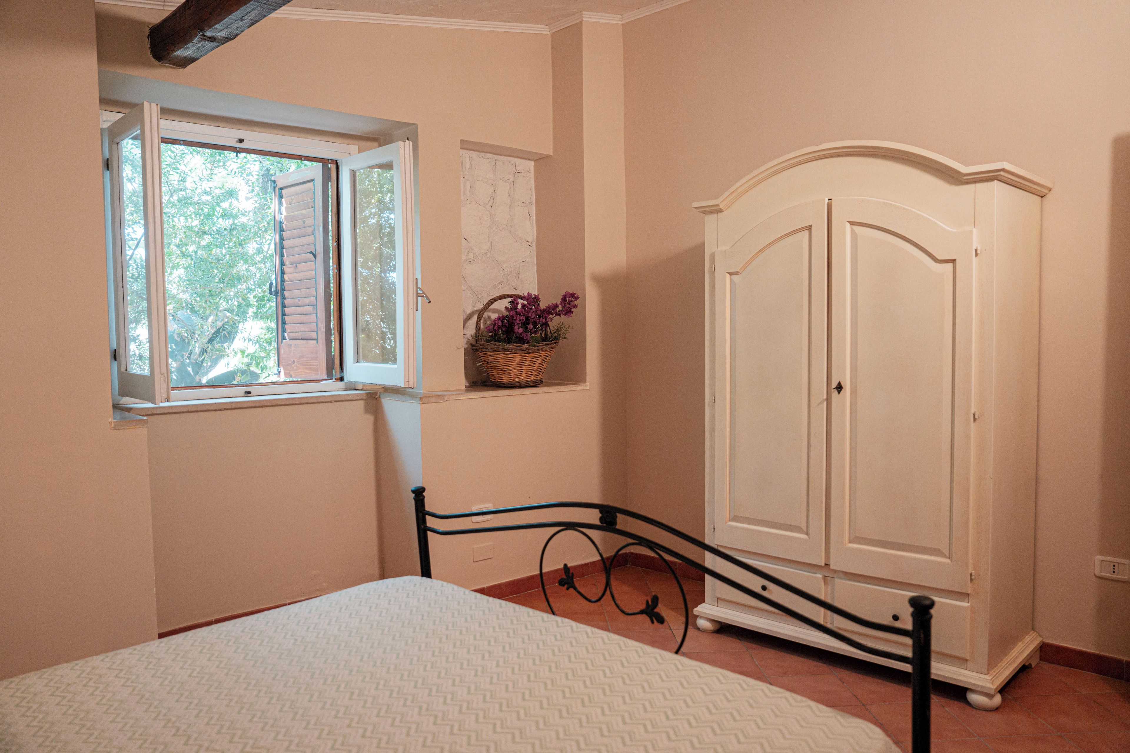 A bedroom with a bed, a window, a wardrobe, and a small wall-mounted shelf with flowers.