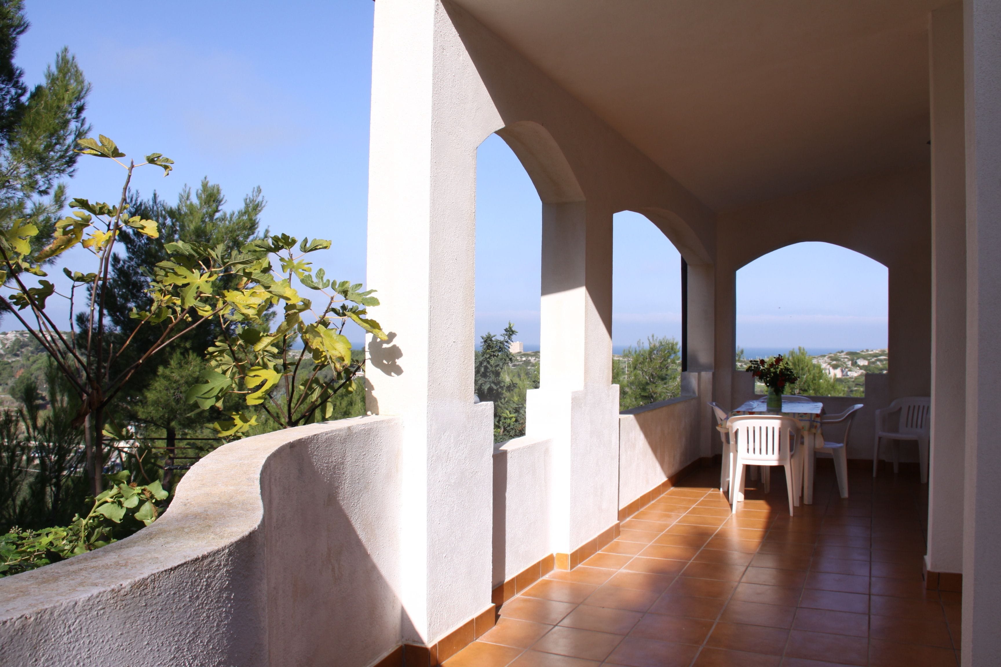 A covered outdoor terrace with white chairs and a table, overlooking a scenic landscape.