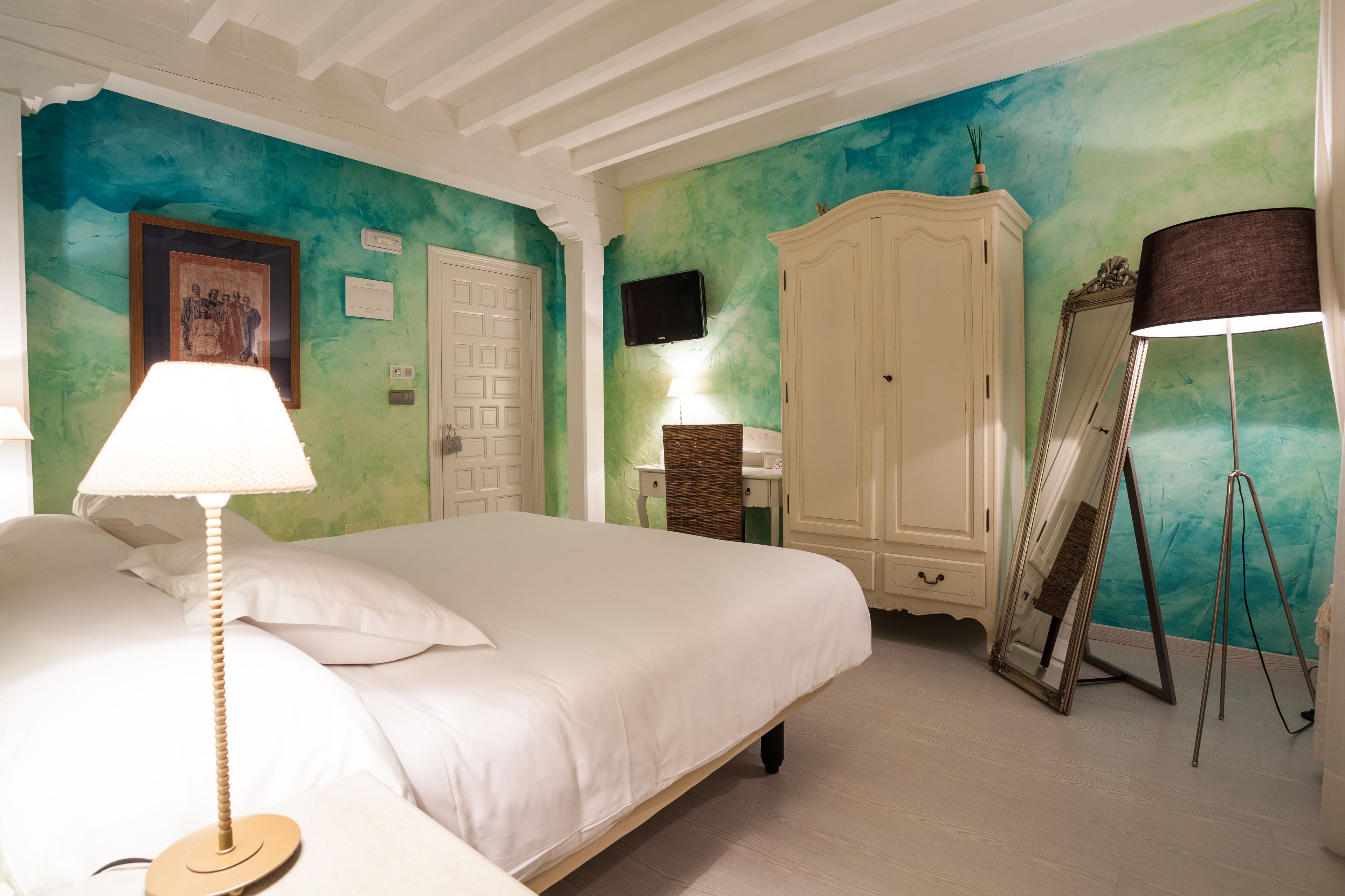 A bedroom with a bed, a bedside lamp, a wardrobe, a television, and a mirror on a stand.