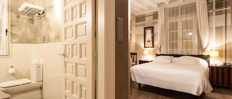 A hotel room with a wooden floor, a bed with white bedding, a nightstand with a lamp, a window with sheer curtains, and a bathroom with a toilet and towel rack.