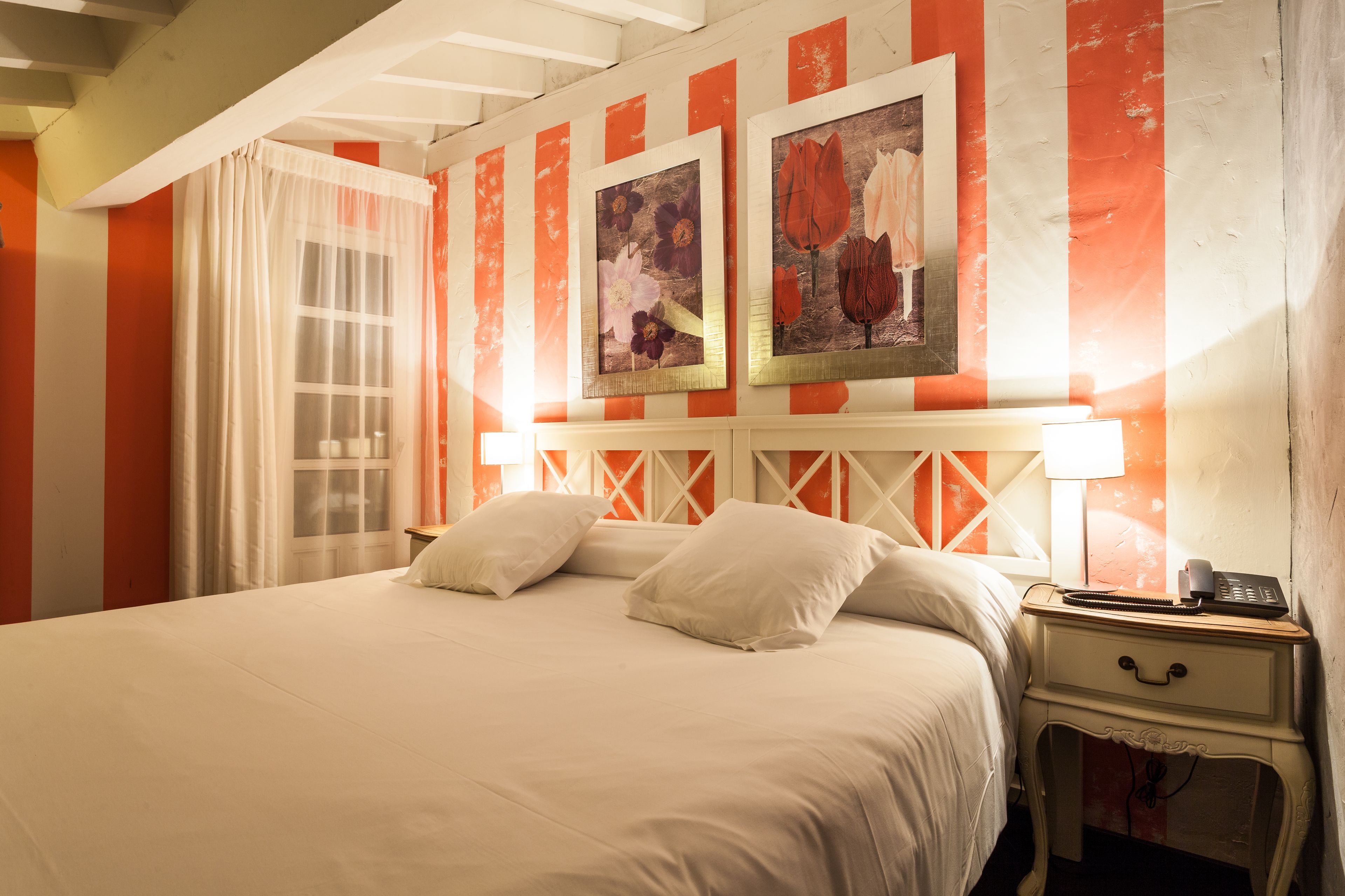 Una cama con ropa blanca, una mesita de noche con lámpara y dos cuadros con flores enmarcados en la pared.