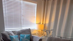 A living room with a grey armchair, a sofa with blue cushions, a lamp, and a window with blinds. - FALL RATES! East end beach, gulf front complex. Lighthouse, Snowbirds welcome. (Dauphin Island)