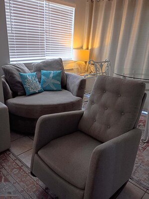 A living room with a grey armchair, a sofa with blue cushions, a lamp, and a window with blinds.