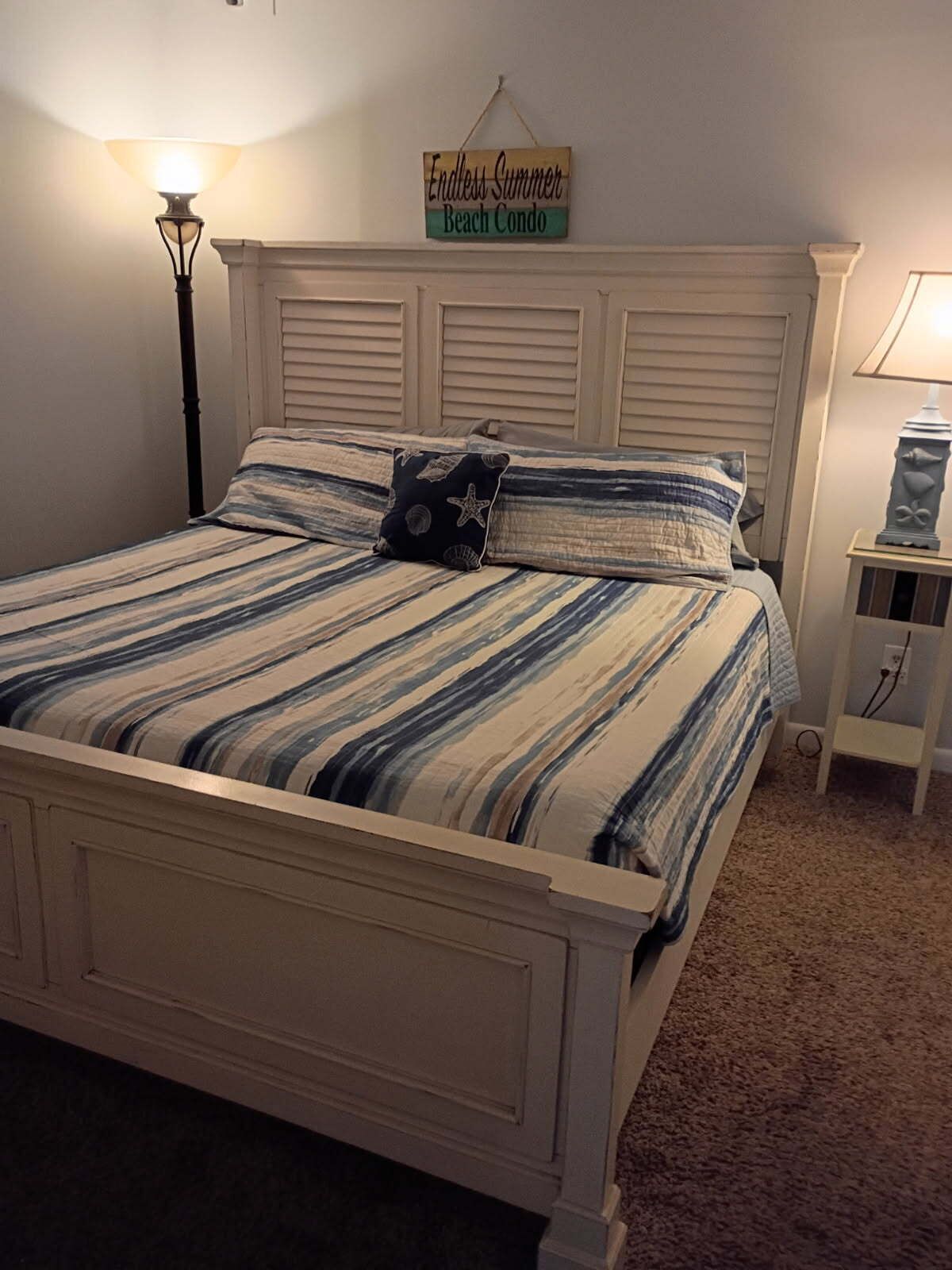 Un lit avec une literie à rayures, une tête de lit, une lampe de chevet, une petite table avec une lampe et un panneau au-dessus du lit.