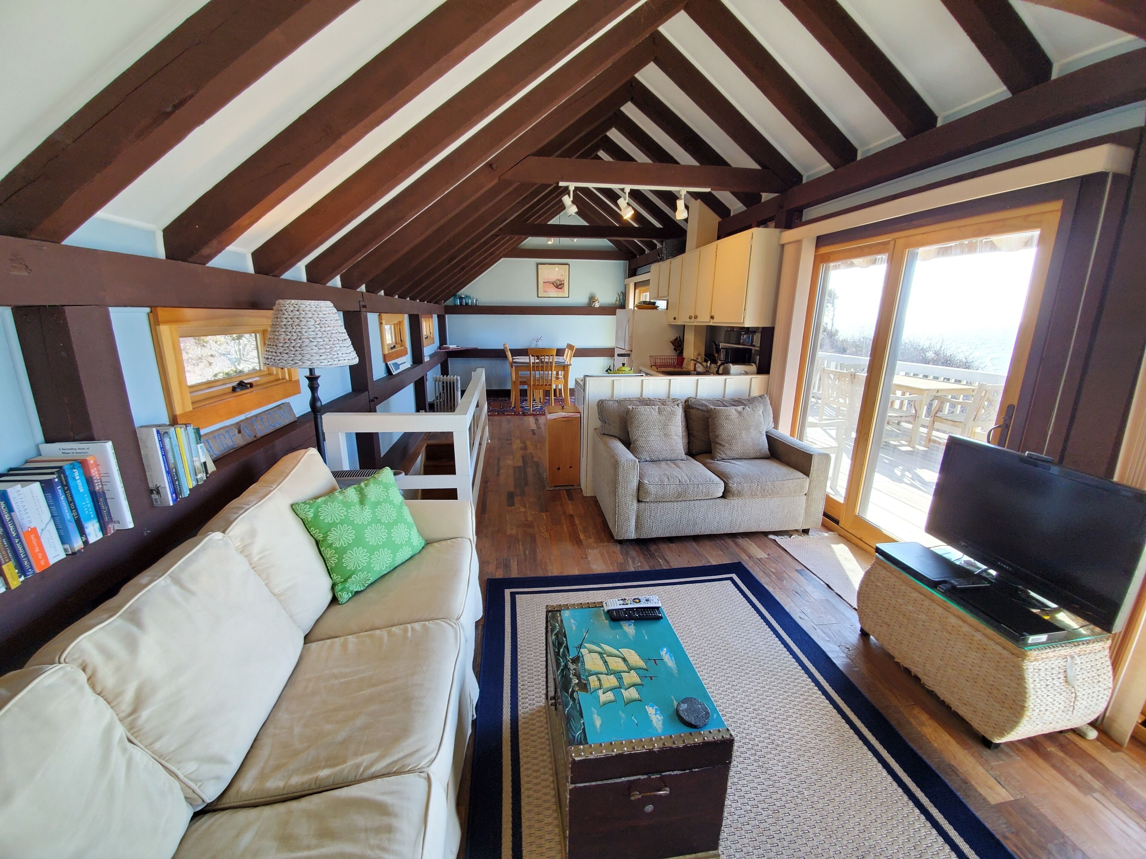A cozy living room with a sofa, a coffee table, and a television. There is a kitchen area in the background with a dining table and chairs.