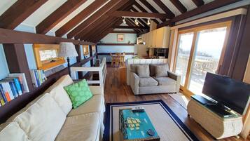 A cozy living room with a sofa, a coffee table, and a television. There is a kitchen area in the background with a dining table and chairs.