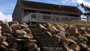A building with a balcony, surrounded by large rocks and a sandy foreground.