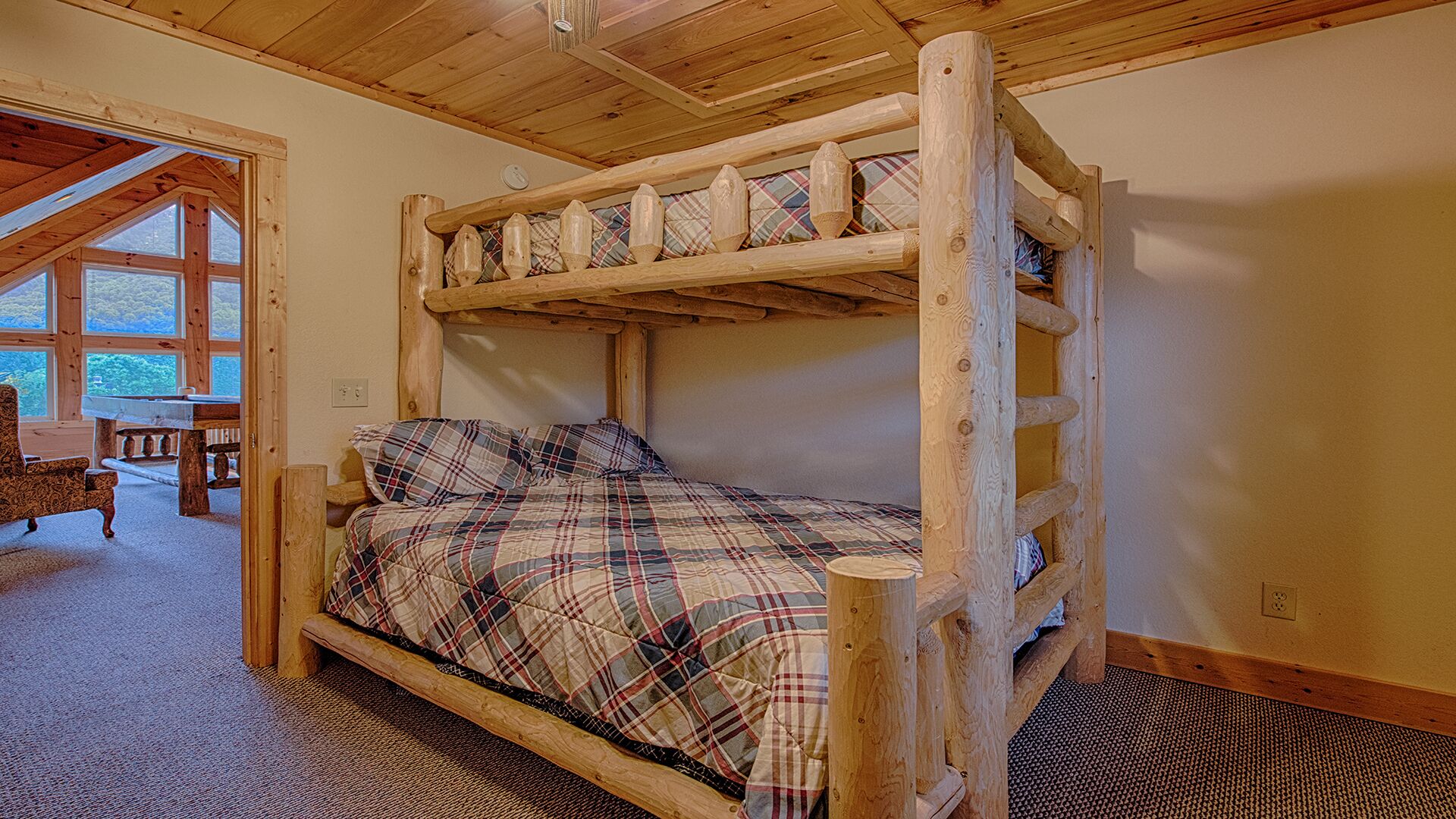 A bunk bed with a plaid bedspread, wooden headboard, and a view of the outdoors through large windows.