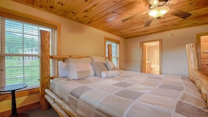 Une chambre avec un grand lit, une tête de lit en bois et un ventilateur de plafond. Il y a une fenêtre avec des stores et une vue sur l’extérieur.