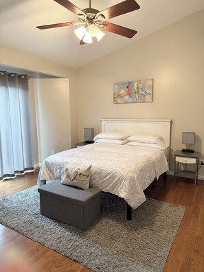 Quarto com cama, mesas de cabeceira, ventilador de teto, pintura e janela com cortinas.
