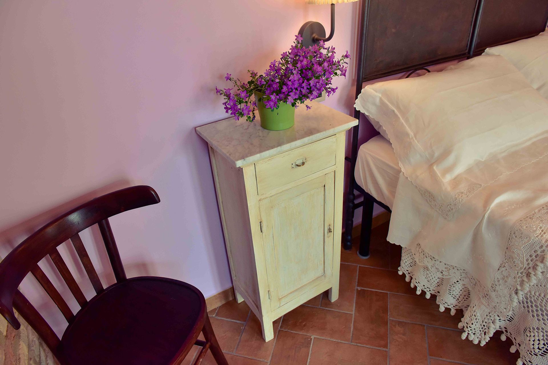 A small, single-bed room with a wooden chair, a nightstand with a green vase of purple flowers, and a white bedspread with lace detailing.
