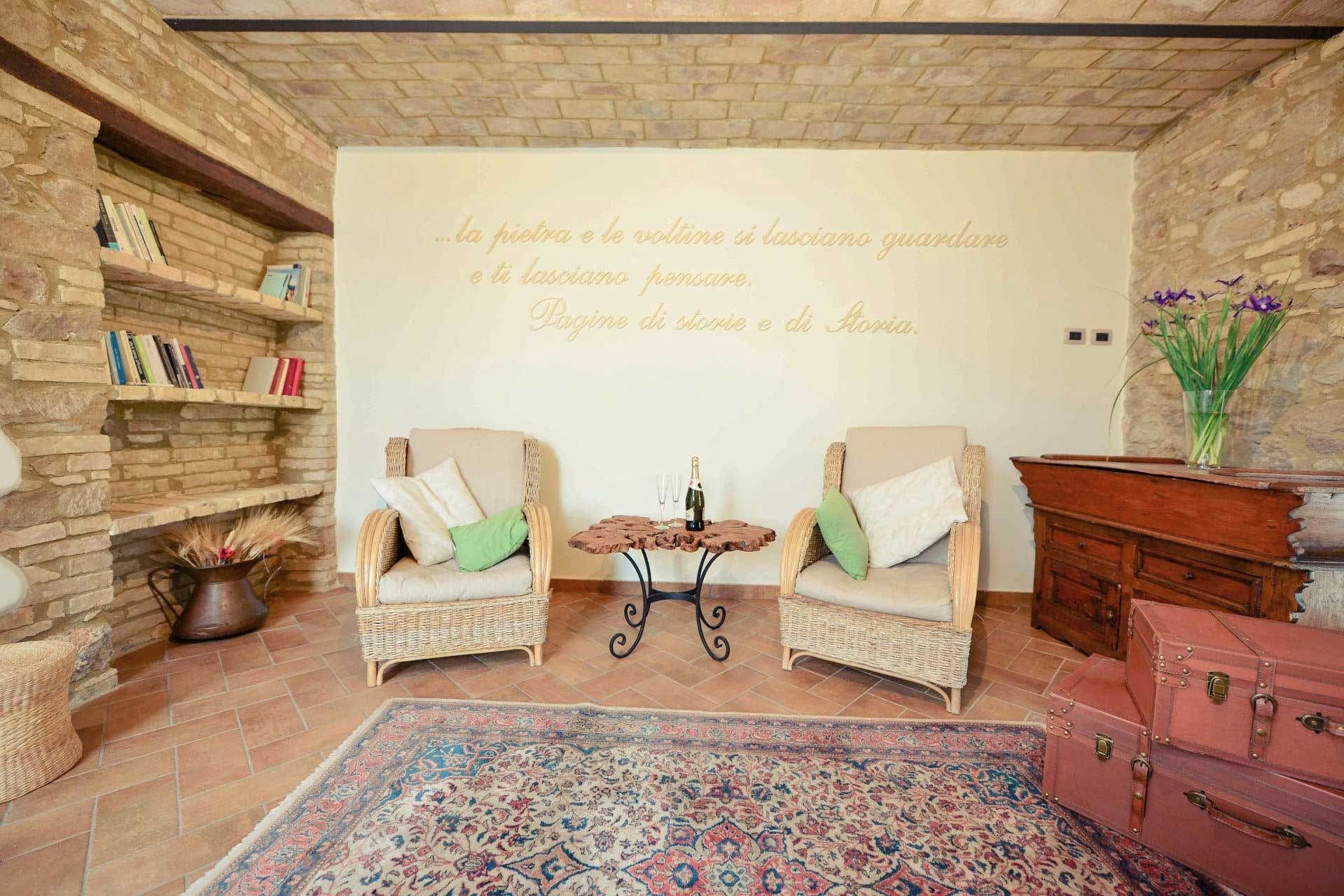 A cozy room with two wicker chairs, a small table with a bottle and glasses, a bookshelf, a decorative wall with a quote, and a patterned rug.