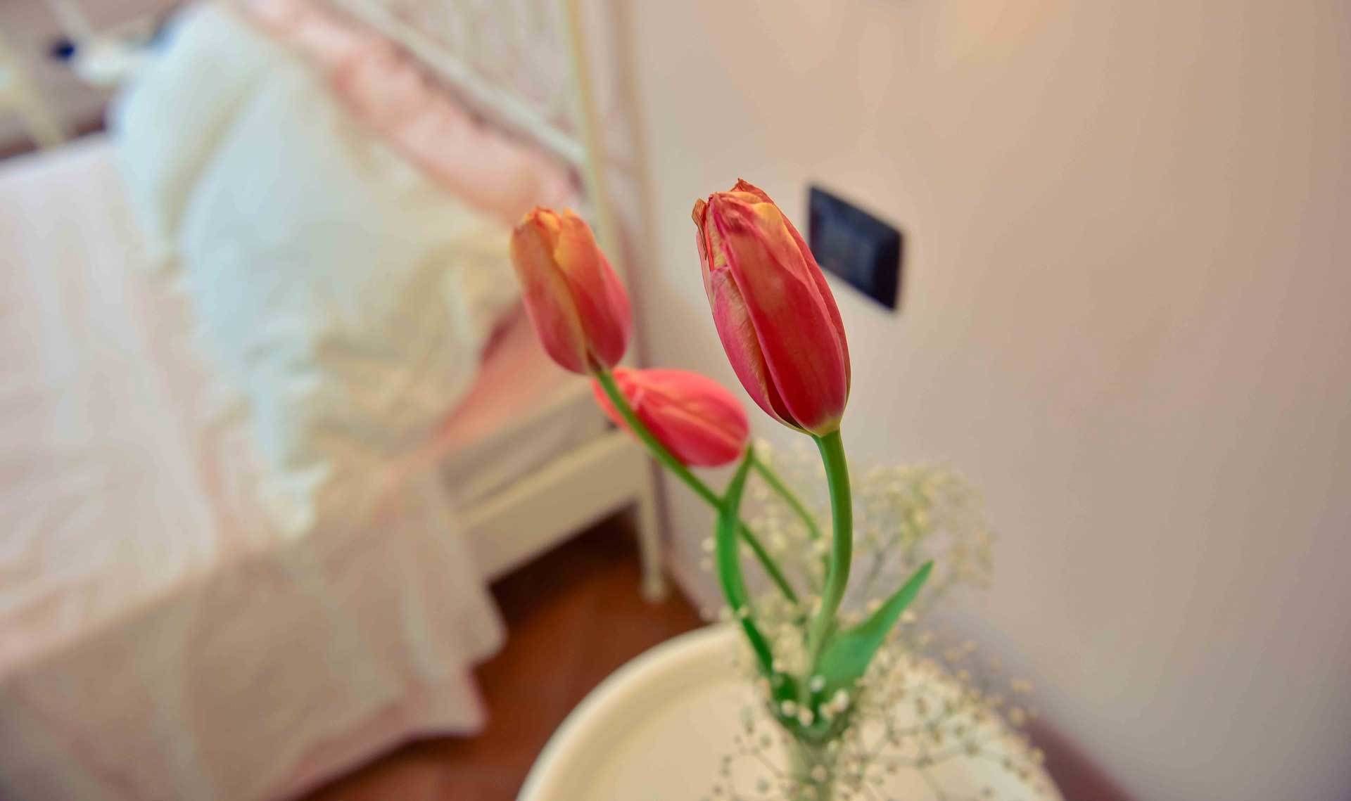 Una habitación con cama, un interruptor de luz en la pared y un jarrón blanco con flores.