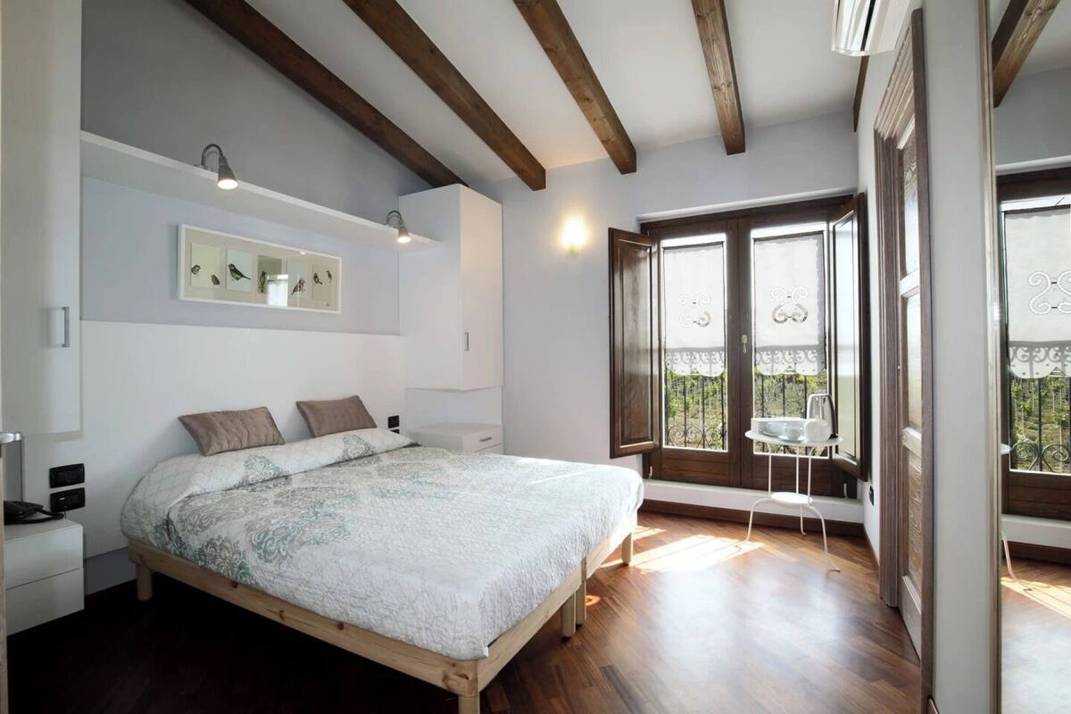 A bedroom with a bed, bedside table, lamp, wall art, and a window with a balcony.