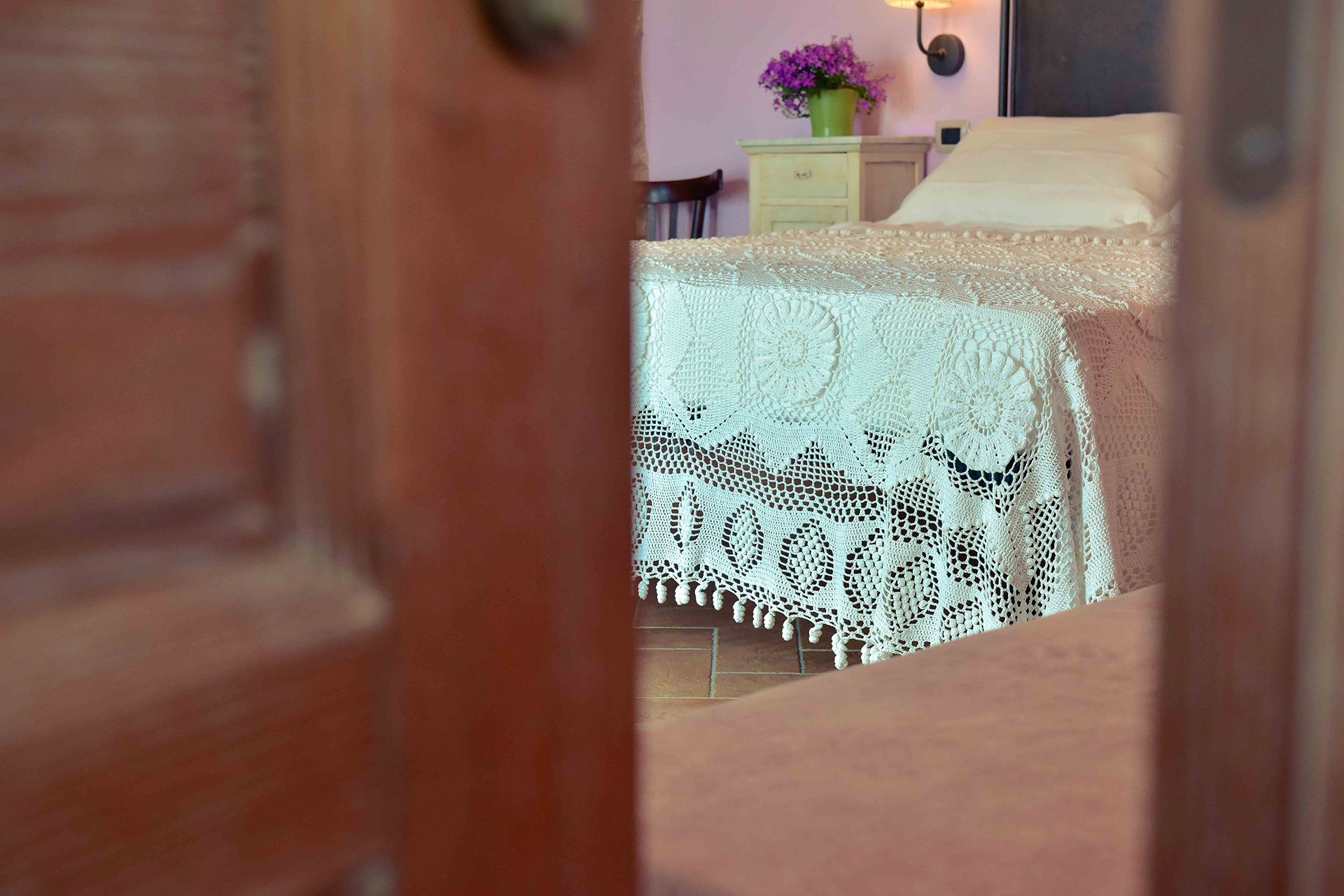 A room with a bed covered in white lace, a wooden door frame, and a small table with a vase of flowers.