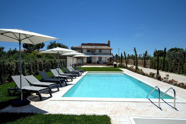A swimming pool with lounge chairs and umbrellas, a house in the background, and a clear sky. - Casale905 (Crecchio)