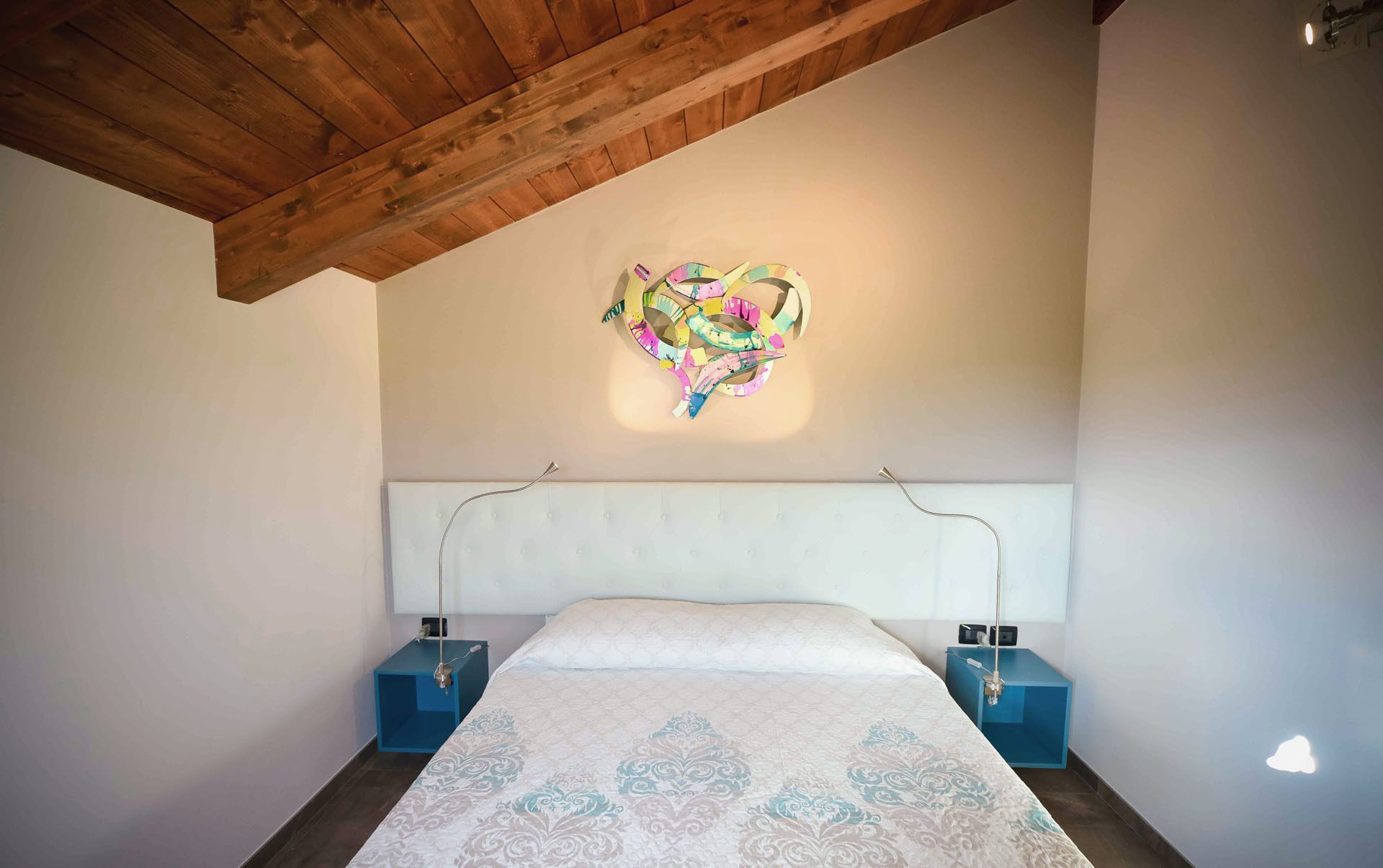 A bedroom with a wooden ceiling, a white headboard, a patterned bedspread, and two bedside tables with lamps.