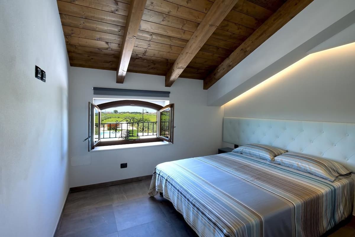 A bedroom with a bed, a window with a view of a green landscape, and a wooden ceiling.