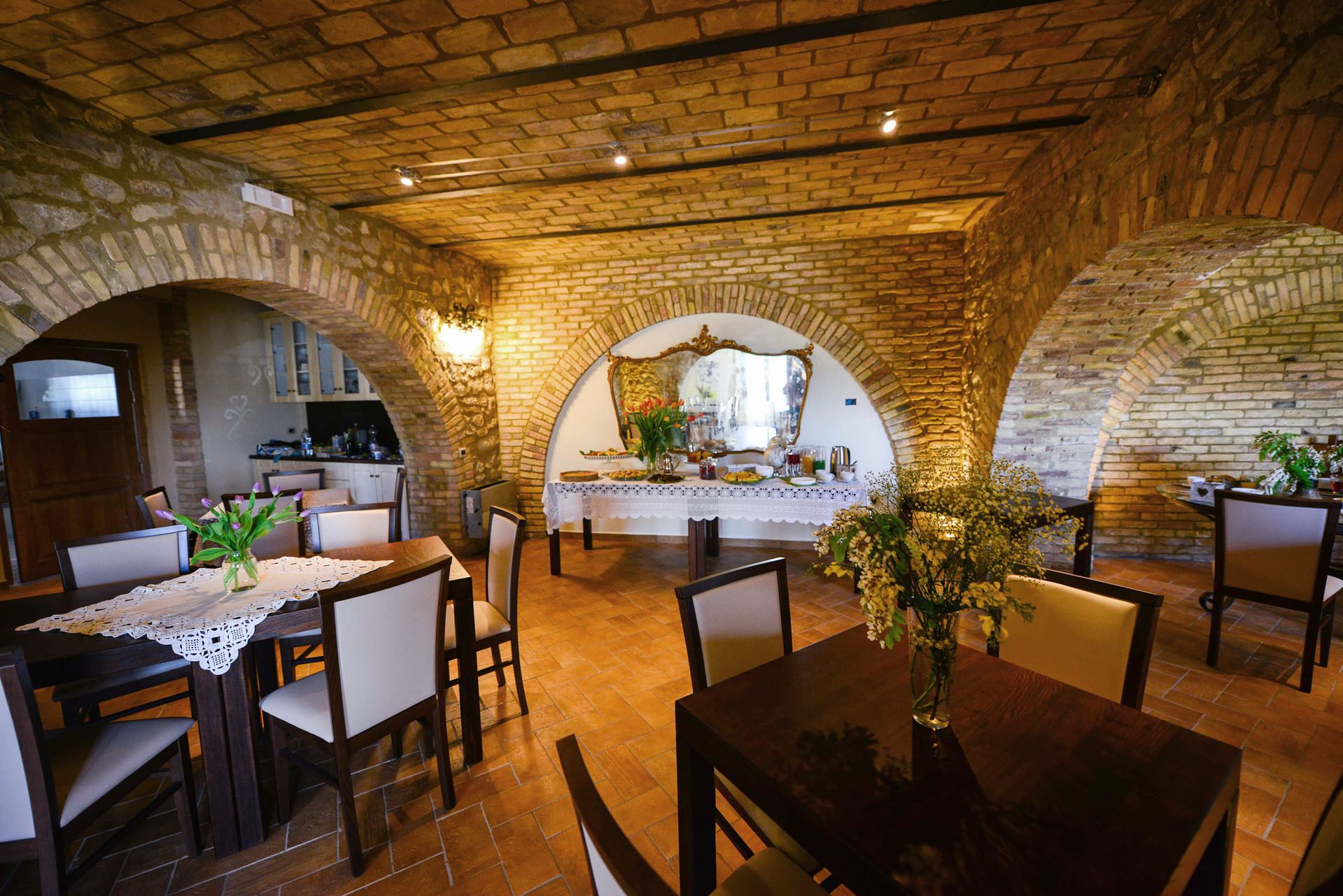 A restaurant with brick arches, wooden tables, and chairs. There is a counter with a mirror and flowers.