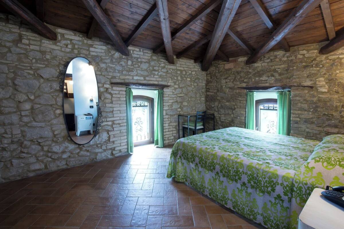 A bedroom with a stone wall, wooden beams, a bed with a green and white bedspread, a mirror, and a small table with a chair.