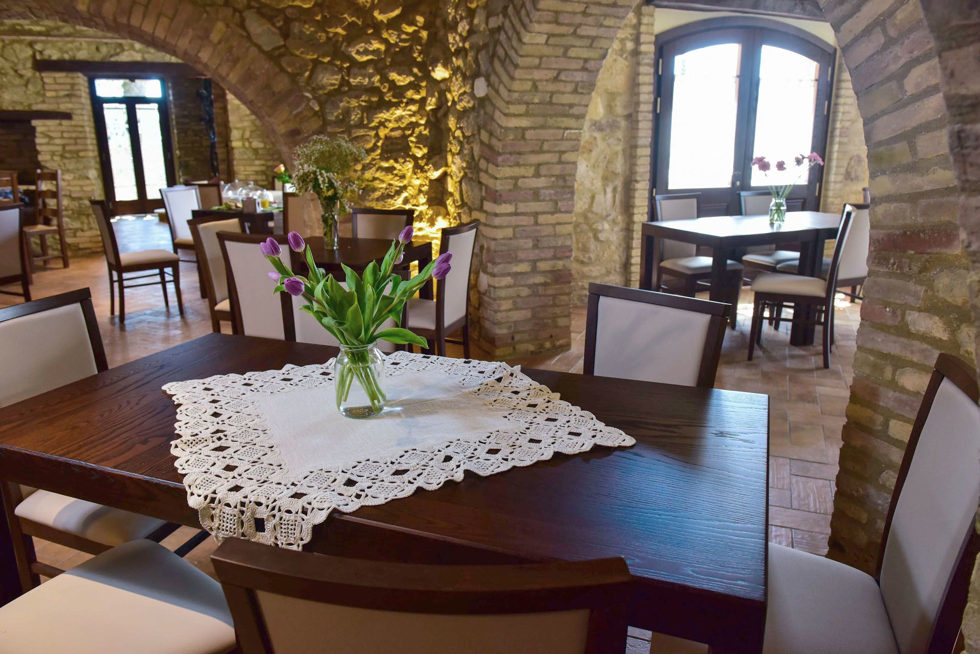 A dining area with a wooden table, white lace tablecloth, and a vase with purple flowers. The room has stone walls and large windows.