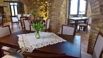 A dining area with a wooden table, white lace tablecloth, and a vase with purple flowers. The room has stone walls and large windows.