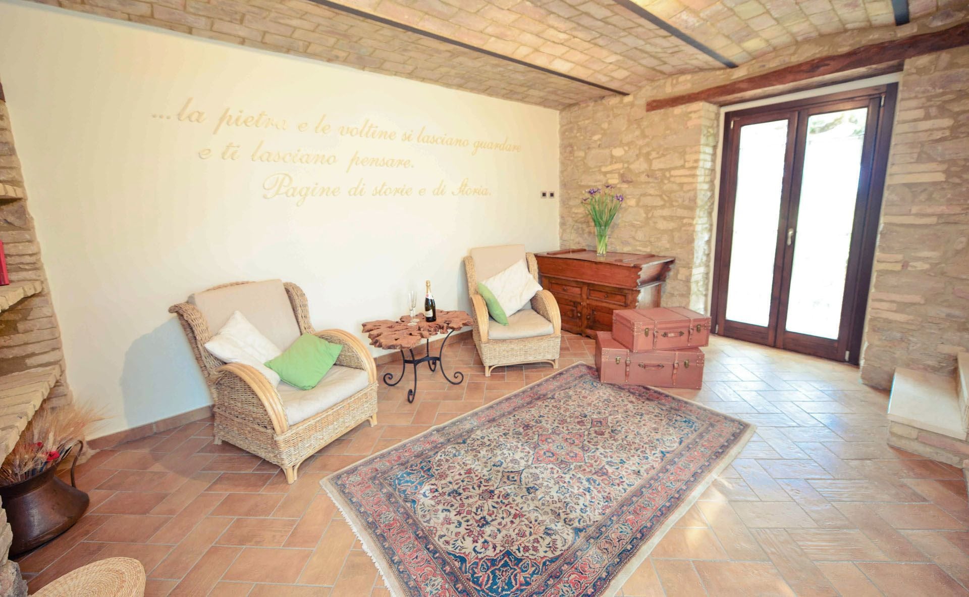 A cozy living room with two wicker chairs, a small table with a bottle, a rug, and a stone wall with a quote.